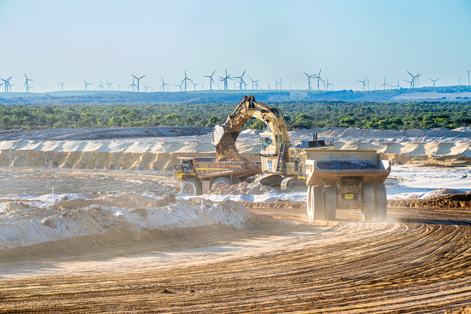 Excavator loading a dump truck at a construction site, with wind turbines visible on the horizon and a clear blue sky.