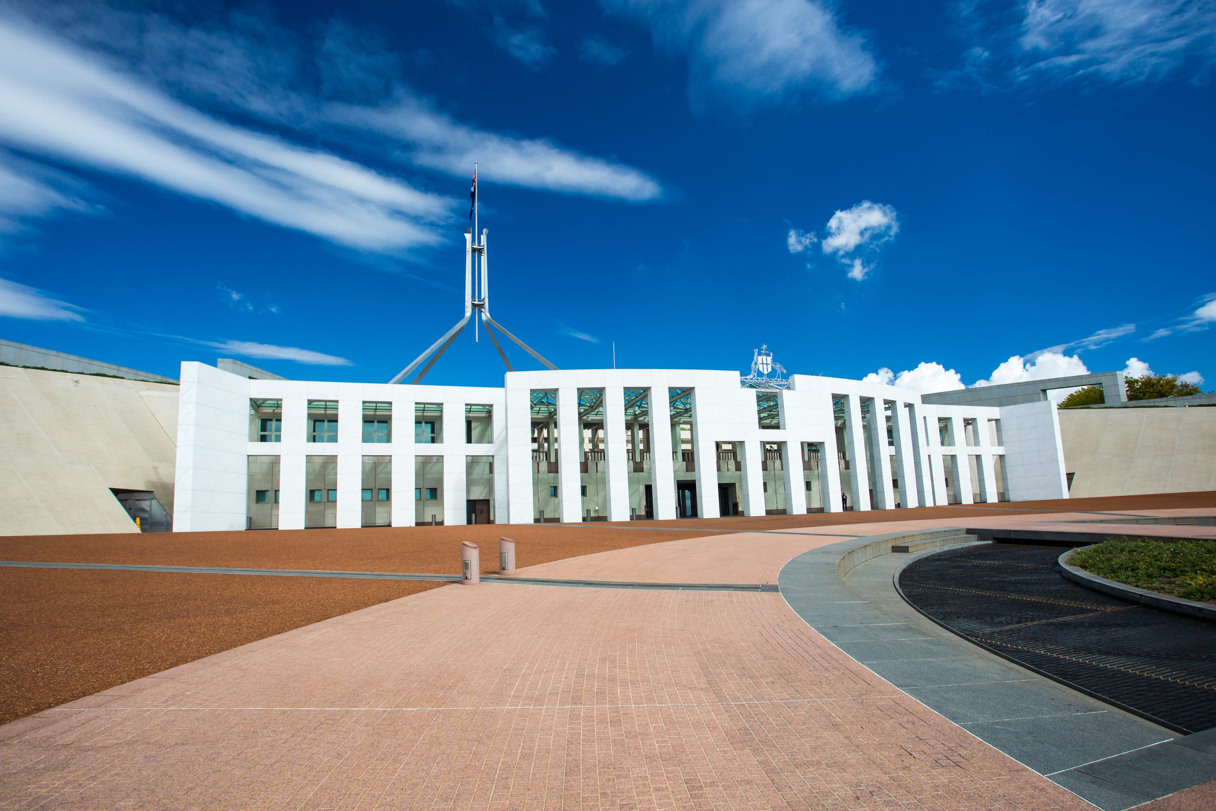 Parliament House in Canberra under a clear blue sky, with its distinct flagpole visible against a backdrop of dramatic clouds.