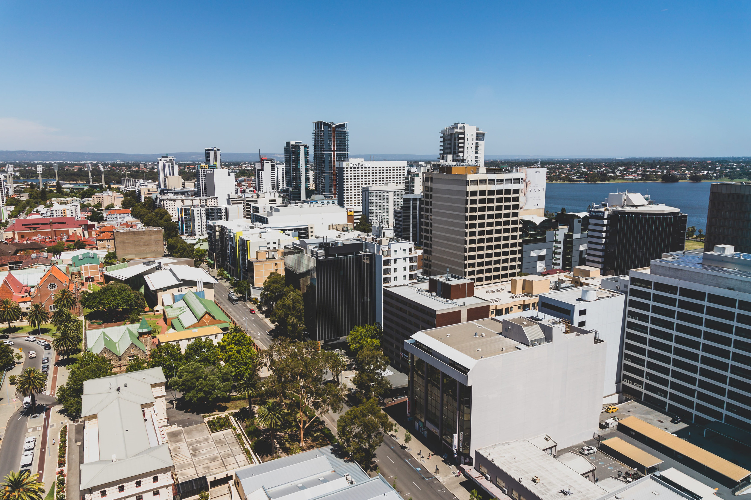 Aerial view of a cityscape with modern high-rise buildings, streets, and greenery, adjacent to a body of water under a clear blue sky.