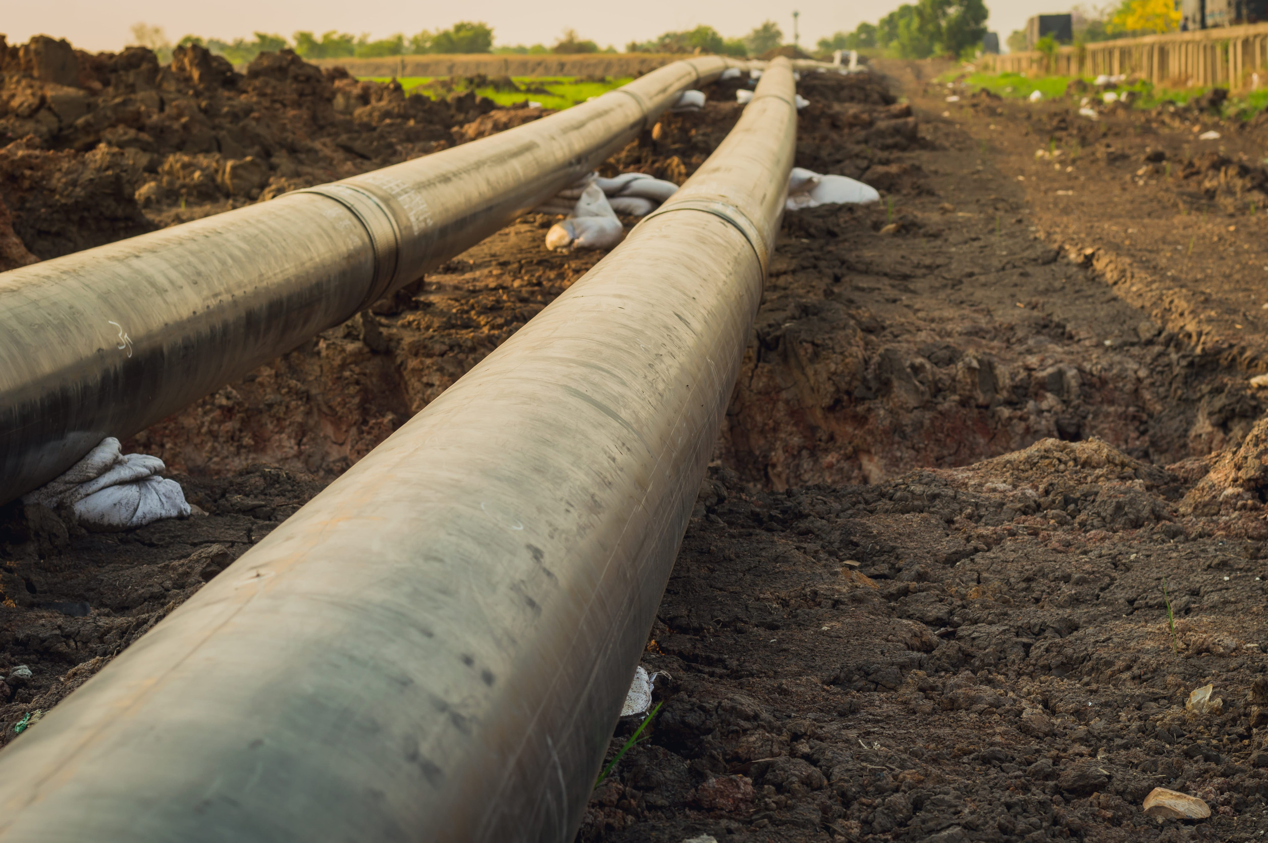 Two large pipelines laid in a trench in a rural area, surrounded by soil and green fields in the background.
