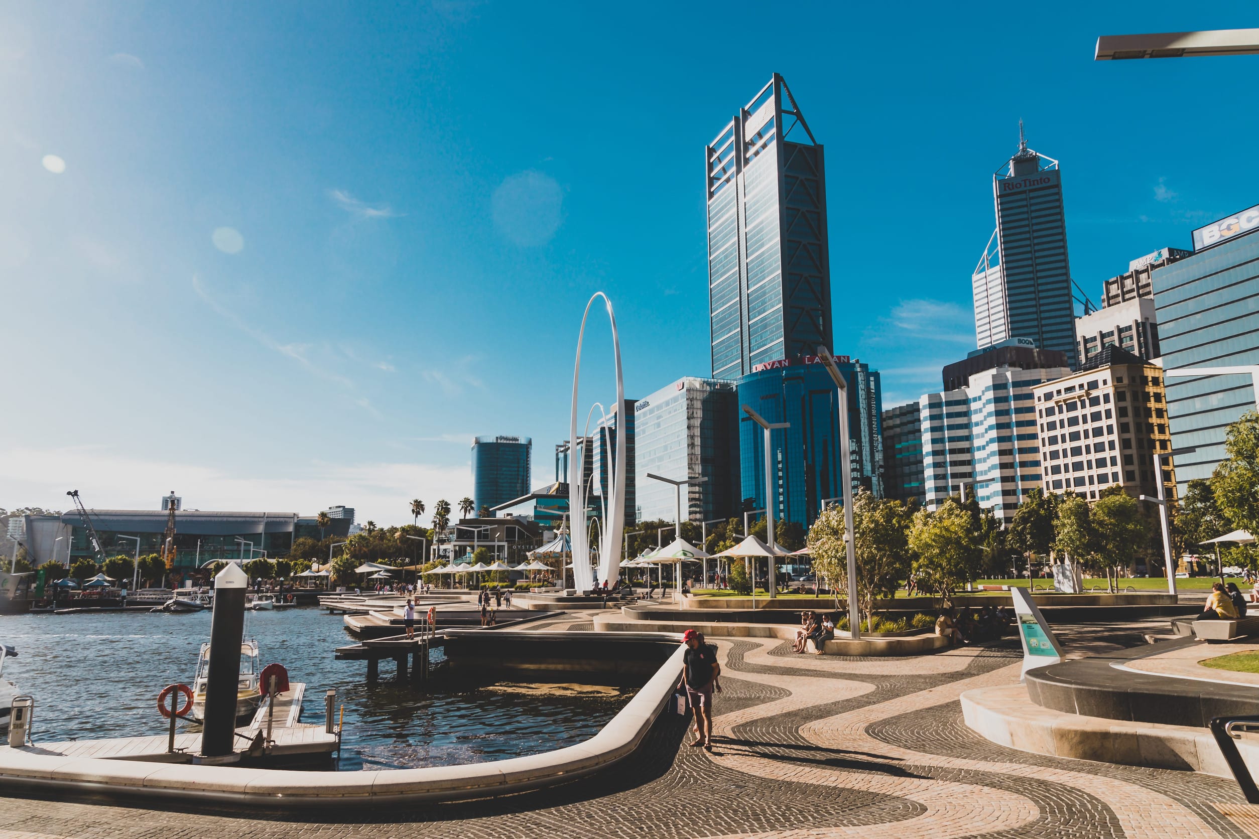 Urban waterfront with modern skyscrapers, curved pathways, and a fountain under a clear blue sky. Pedestrians stroll along the promenade.