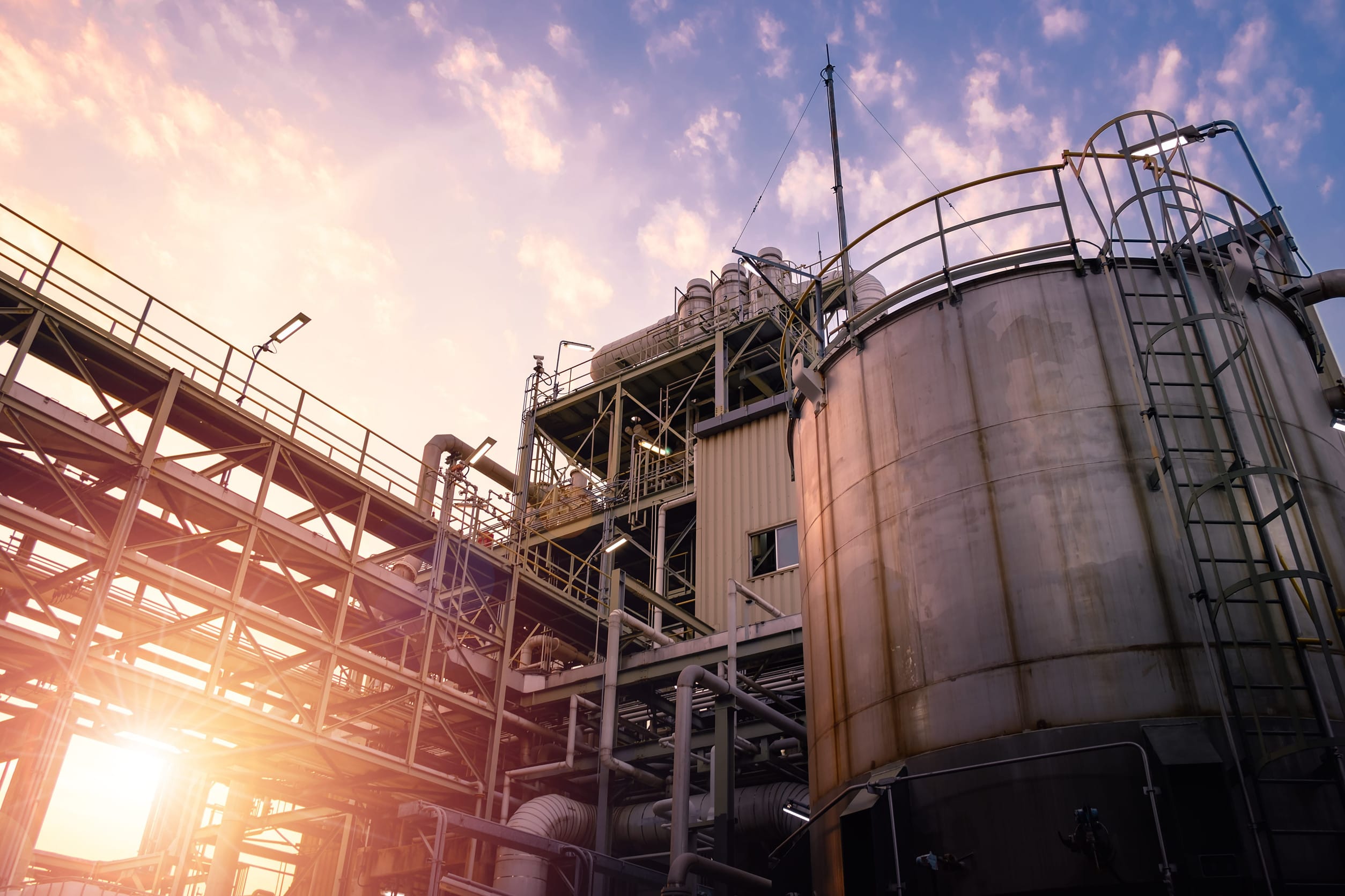 Industrial facility with pipes and tanks at sunset, bathed in warm light. Metal structures and ladders are visible against a colorful sky.