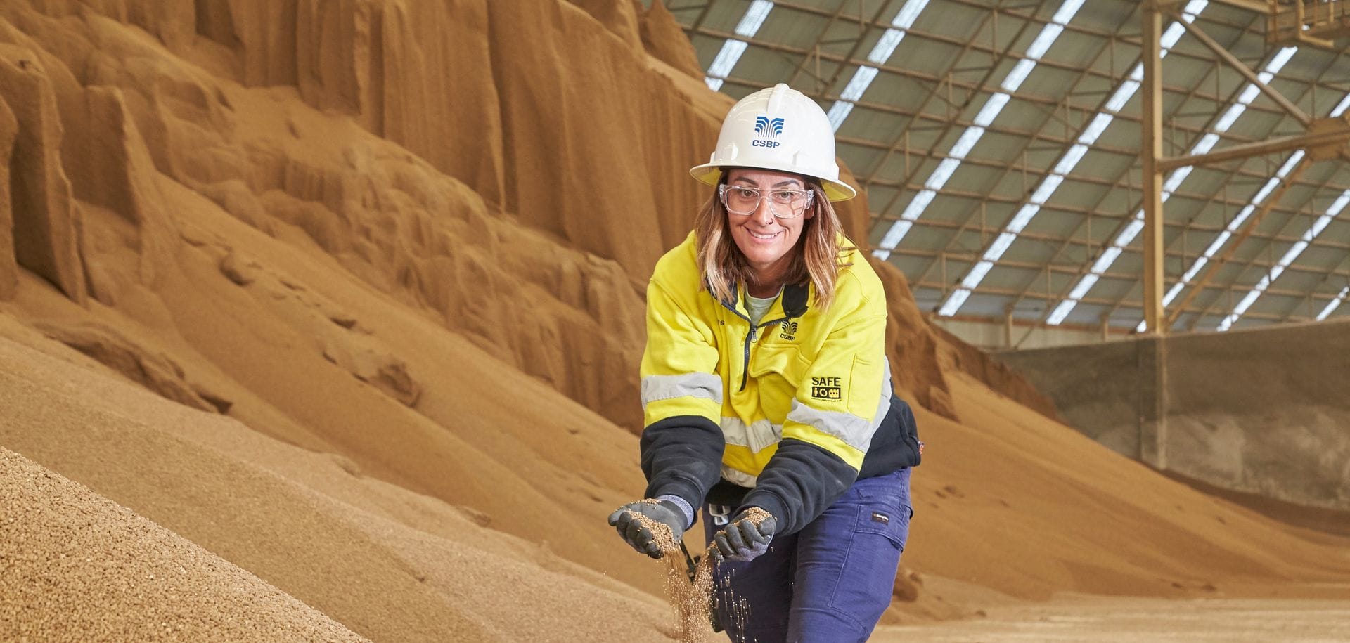 A person in a hard hat and hi-vis jacket handling a pile of brown granules inside a large storage facility.
