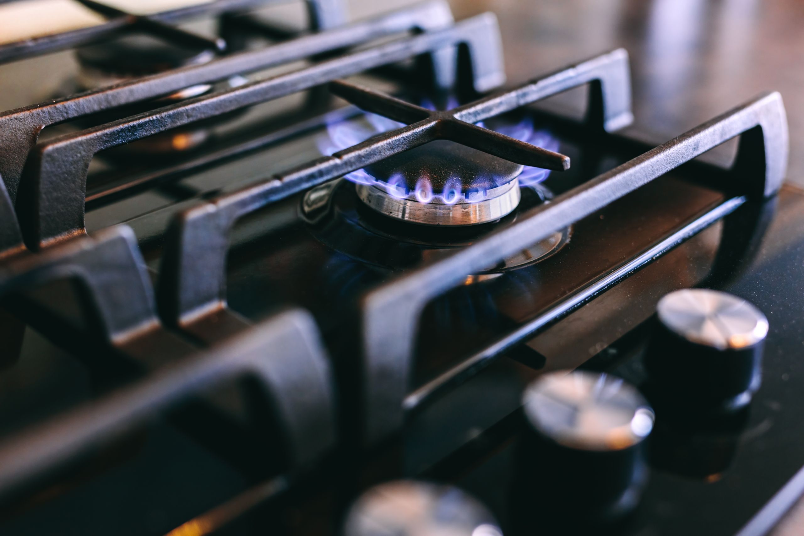 Close-up of a lit gas stove burner with blue flame, surrounded by black metal grates, and control knobs on the right.