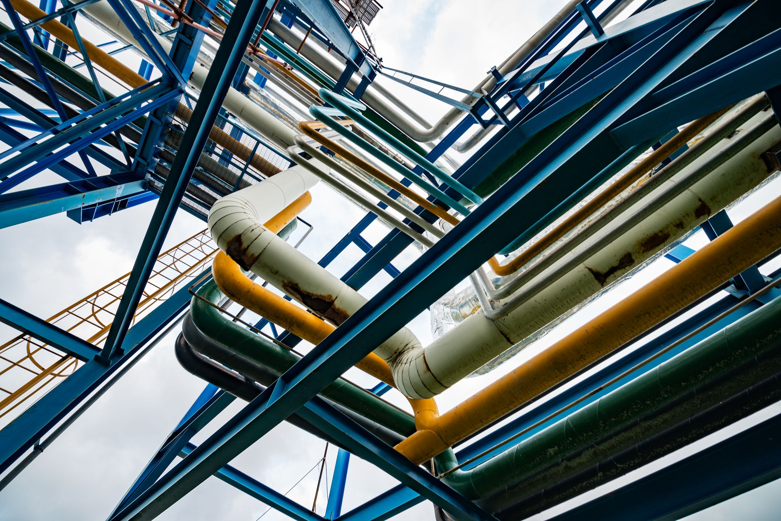 Complex network of colorful industrial pipes and beams against a cloudy sky, viewed from a low angle.