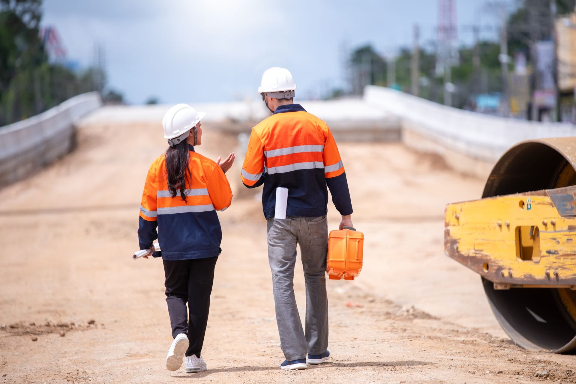 Two construction workers in orange safety jackets and helmets walk on a dirt road, carrying tools, with machinery on the side.